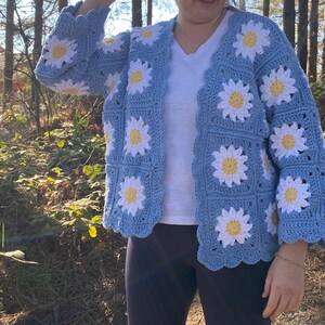 K&ouml;nnte beinhalten: Eine hellblaue geh&auml;kelte Strickjacke mit einem wei&szlig;en G&auml;nsebl&uuml;mchenmuster. Die Strickjacke hat einen V-Ausschnitt und lange &Auml;rmel.