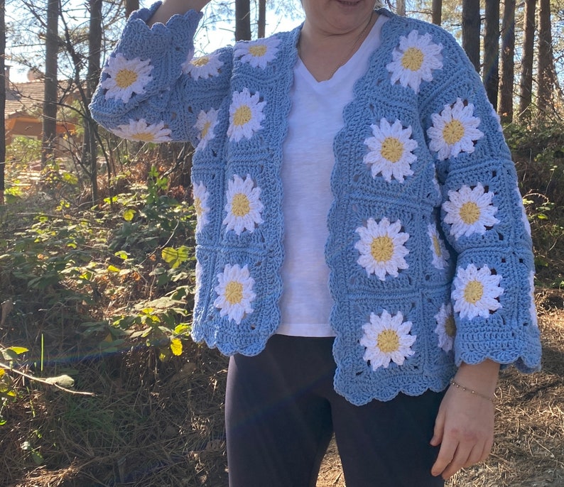 K&ouml;nnte beinhalten: Eine hellblaue geh&auml;kelte Strickjacke mit einem wei&szlig;en G&auml;nsebl&uuml;mchenmuster. Die Strickjacke hat einen V-Ausschnitt und lange &Auml;rmel.