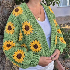 May include: Green crochet cardigan with sunflower motifs. The cardigan has a V-neck and long sleeves.