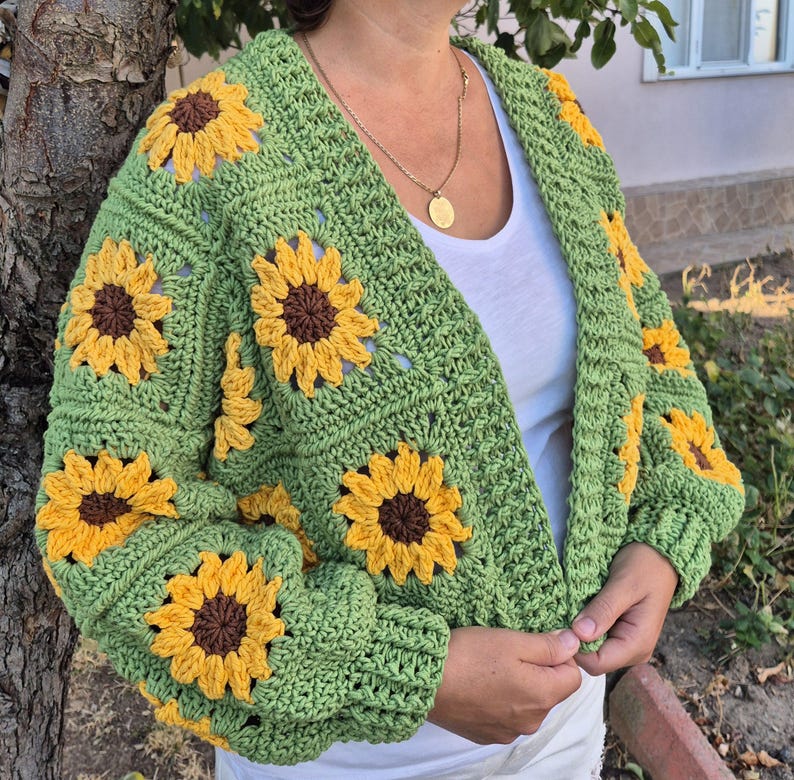 May include: Green crochet cardigan with sunflower motifs. The cardigan has a V-neck and long sleeves.
