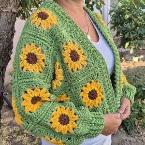 May include: A green crocheted cardigan with a sunflower pattern. The cardigan has a V-neck and long sleeves.