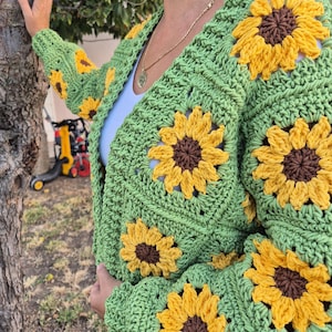 May include: Green crochet cardigan with sunflower motifs. The cardigan has a V-neck and long sleeves.