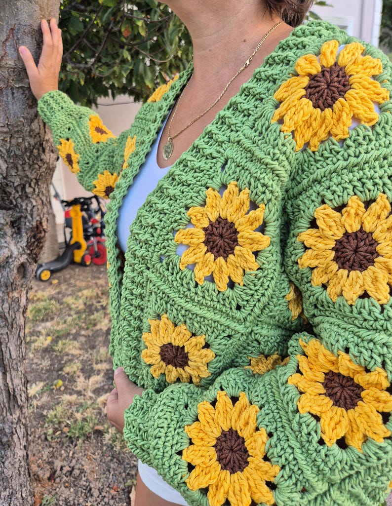 May include: Green crochet cardigan with sunflower motifs. The cardigan has a V-neck and long sleeves.