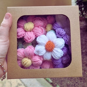 Puede incluir: Un conjunto de seis flores de crochet en una caja de cartón. Las flores son en tonos de rosa, blanco y morado. Las flores están dispuestas en un ramo.
