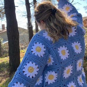 K&ouml;nnte beinhalten: Eine hellblaue geh&auml;kelte Strickjacke mit wei&szlig;en G&auml;nsebl&uuml;mchen. Die Strickjacke hat einen gewellten Saum und lange &Auml;rmel.
