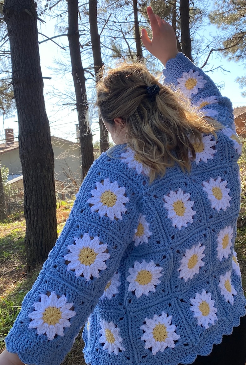 K&ouml;nnte beinhalten: Eine hellblaue geh&auml;kelte Strickjacke mit wei&szlig;en G&auml;nsebl&uuml;mchen. Die Strickjacke hat einen gewellten Saum und lange &Auml;rmel.