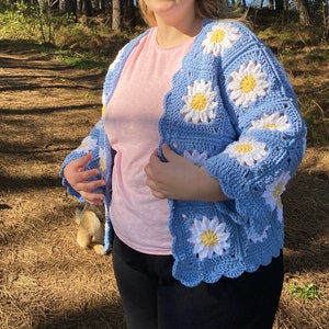 K&ouml;nnte beinhalten: Eine blaue geh&auml;kelte Strickjacke mit einem G&auml;nsebl&uuml;mchenmuster. Die Strickjacke hat eine lockere Passform und ist aus weichem Garn gefertigt.