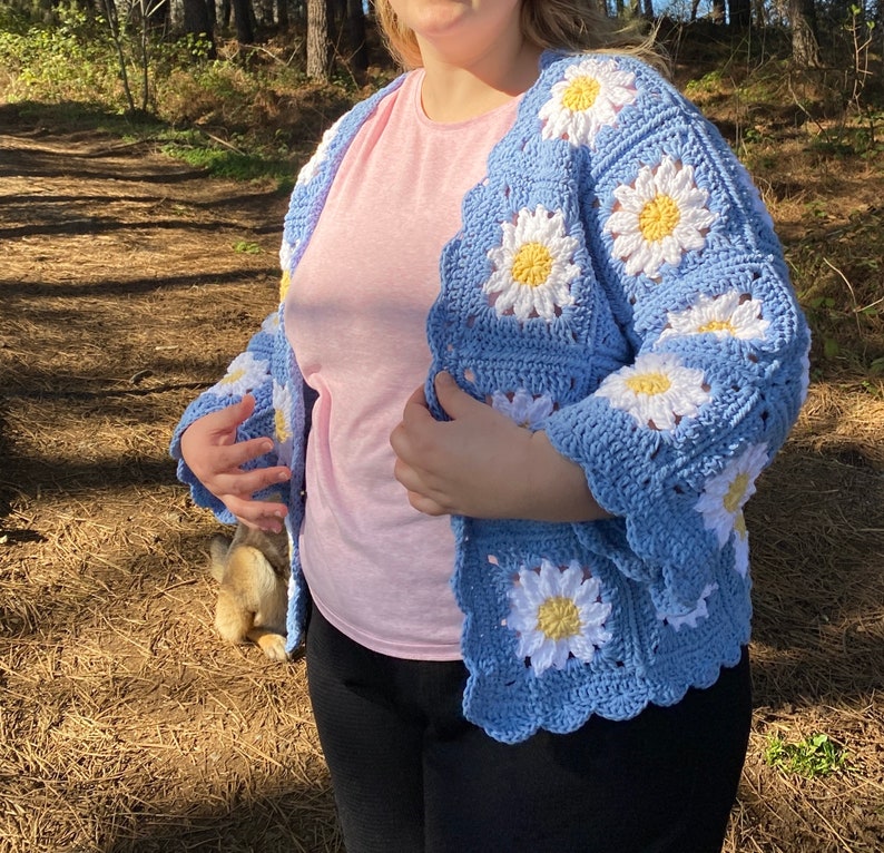 K&ouml;nnte beinhalten: Eine blaue geh&auml;kelte Strickjacke mit einem G&auml;nsebl&uuml;mchenmuster. Die Strickjacke hat eine lockere Passform und ist aus weichem Garn gefertigt.