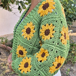 May include: Green crocheted cardigan with sunflower motifs. The cardigan has a V-neck and long sleeves.