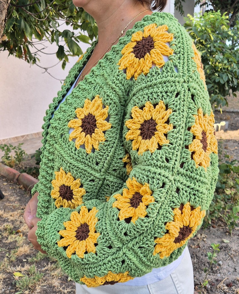 May include: Green crocheted cardigan with sunflower motifs. The cardigan has a V-neck and long sleeves.