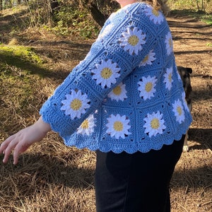 K&ouml;nnte beinhalten: Eine hellblaue geh&auml;kelte Strickjacke mit einem G&auml;nsebl&uuml;mchenmuster. Die Strickjacke hat einen gewellten Saum und Dreiviertel&auml;rmel.