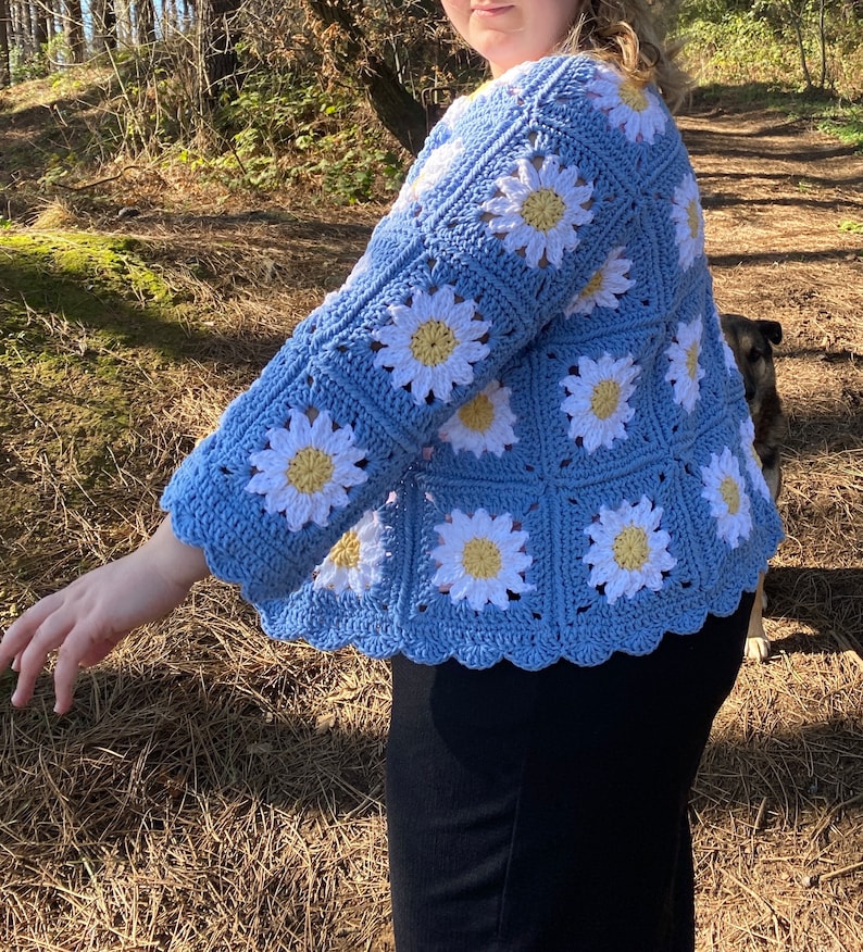 K&ouml;nnte beinhalten: Eine hellblaue geh&auml;kelte Strickjacke mit einem G&auml;nsebl&uuml;mchenmuster. Die Strickjacke hat einen gewellten Saum und Dreiviertel&auml;rmel.
