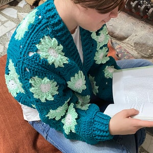 May include: A teal blue crocheted cardigan with green and white flowers. The cardigan is worn by a person sitting on a brown rug and reading a book.