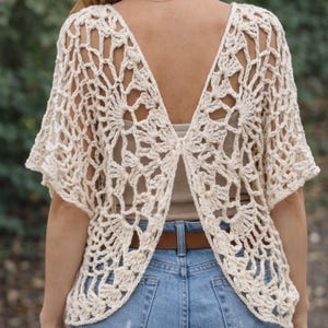 May include: Cream-colored, open-back crochet top with short sleeves. The intricate floral pattern is visible. The top is paired with blue jeans and a brown belt. The back of the top is open, revealing the wearer's back.