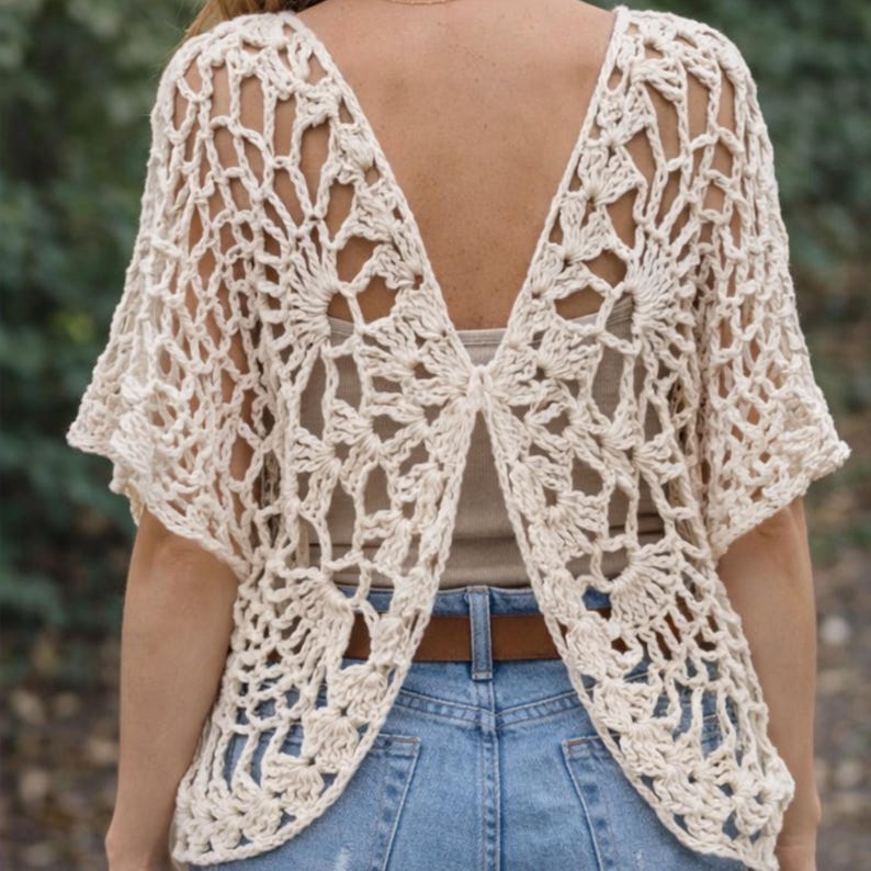 May include: Cream-colored, open-back crochet top with short sleeves. The intricate floral pattern is visible. The top is paired with blue jeans and a brown belt. The back of the top is open, revealing the wearer's back.