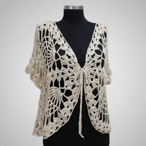 May include: A cream-colored, open-front crochet shawl with a V-neck and short sleeves. The shawl features a delicate, openwork design and a tie closure at the bust. The garment is displayed on a black mannequin.