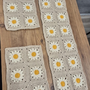May include: Handmade crochet daisy table runner and two matching placemats. The daisy flowers have a yellow center and white petals, set against a beige background. The items are arranged on a wooden surface.