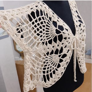 May include: A cream-colored, open-front crochet shrug with a delicate, openwork design. The shrug features a tie closure at the front and flared sleeves. The intricate pattern and neutral color make it a versatile layering piece.
