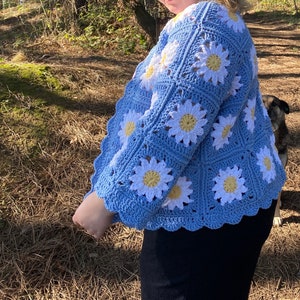 K&ouml;nnte beinhalten: Eine hellblaue geh&auml;kelte Strickjacke mit einem G&auml;nsebl&uuml;mchenmuster. Die Strickjacke hat einen gewellten Saum und ist aus wei&szlig;em und gelbem Garn gefertigt.