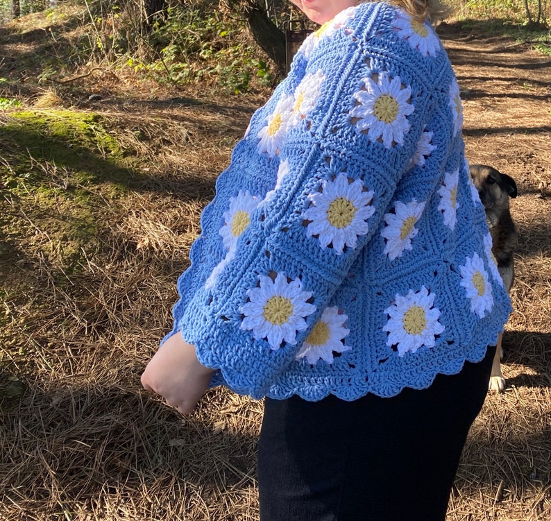 K&ouml;nnte beinhalten: Eine hellblaue geh&auml;kelte Strickjacke mit einem G&auml;nsebl&uuml;mchenmuster. Die Strickjacke hat einen gewellten Saum und ist aus wei&szlig;em und gelbem Garn gefertigt.