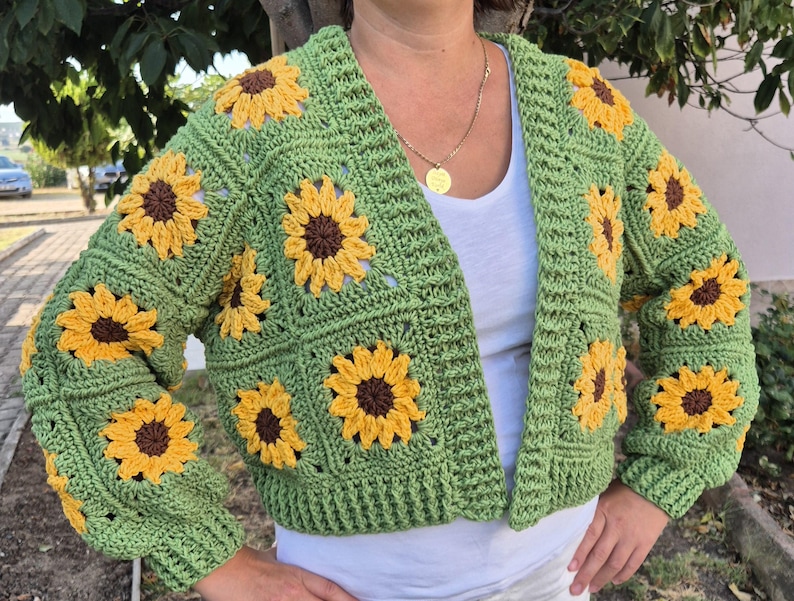 May include: A green crocheted cardigan with a sunflower pattern. The cardigan has a V-neck and long sleeves.