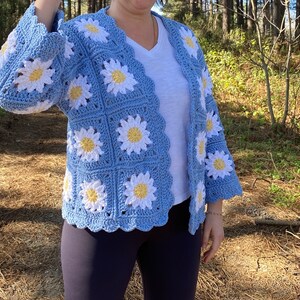 K&ouml;nnte beinhalten: Eine hellblaue geh&auml;kelte Strickjacke mit einem G&auml;nsebl&uuml;mchenmuster. Die Strickjacke hat einen V-Ausschnitt und lange &Auml;rmel.