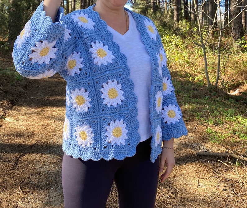 K&ouml;nnte beinhalten: Eine hellblaue geh&auml;kelte Strickjacke mit einem G&auml;nsebl&uuml;mchenmuster. Die Strickjacke hat einen V-Ausschnitt und lange &Auml;rmel.