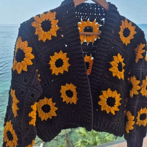 May include: A crochet cardigan featuring a repeating pattern of black and yellow sunflower motifs. The garment is constructed from individual square panels, each showcasing a central black circle surrounded by bright yellow petals, joined together.