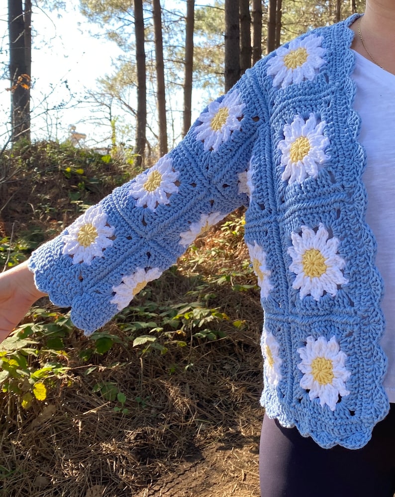 K&ouml;nnte beinhalten: Eine hellblaue geh&auml;kelte Strickjacke mit einem G&auml;nsebl&uuml;mchenmuster. Die Strickjacke hat lange &Auml;rmel und einen gewellten Saum.