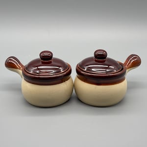 Puede incluir: Dos cuencos de sopa de cerámica con tapas. Los cuencos son de color blanquecino con un borde y asa marrón. Las tapas son marrones con un pequeño pomo en la parte superior. Los cuencos son redondos y tienen un asa.