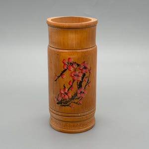 Puede incluir: Un recipiente cilíndrico de bambú con un acabado de madera natural. El recipiente presenta un diseño floral pintado con flores rosas y rojas. El recipiente se utiliza probablemente para guardar lápices, pinceles u otros objetos pequeños.