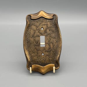 May include: A bronze-colored light switch cover with a textured, organic pattern. The cover has a single switch opening and two screw holes. The design features a decorative, shield-like shape with a curved top and bottom.