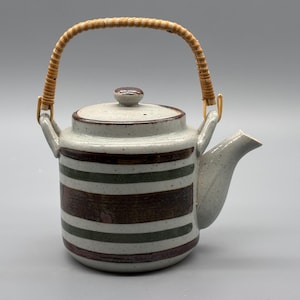 Peut inclure: Une théière en céramique avec un motif à rayures marron, vert et blanc. Elle possède une anse tressée et un bec verseur. La théière est de couleur gris clair avec une finition mouchetée.