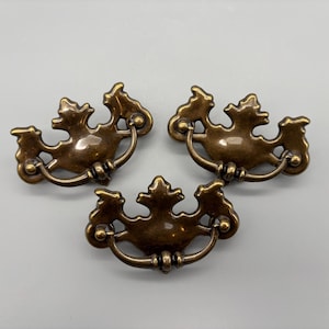 以下が含まれることがあります： 装飾的なフローラルデザインが施された、3つのアンティークブロンズ製の引き出し取っ手。各取っ手は、湾曲したハンドルと装飾的なバックプレートを備えています。取っ手は金属製で、暖かく、使い込まれたような仕上がりで、ヴィンテージ家具の修復に最適です。