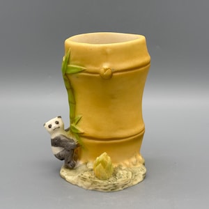 May include: A ceramic vase shaped like a bamboo stalk, painted in shades of yellow and tan. A small panda bear clings to the side, with a bamboo shoot at the base. The vase is approximately 13 cm tall.