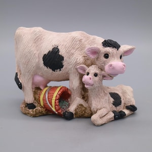 Op de afbeelding: Een beeldje van een koe en een kalf. De koe is lichtroze met zwarte vlekken en een zwarte vlek op haar hoofd. Het kalf is lichtroze met zwarte vlekken en ligt. Een rode en gele emmer staat bij de voeten van de koe.