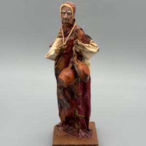 Op de afbeelding: Een handgemaakte papieren mache figuur van een figuur met een kap, met twee bruine kruiken. De figuur is gekleed in bordeauxrode en crèmekleurige gewaden, met een touw om de nek. Het beeldje is op een bruine basis gemonteerd.
