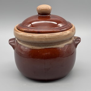 Puede incluir: Una olla de cerámica marrón con tapa y borde y asa de color marrón claro. La olla tiene forma redondeada con dos asas pequeñas a los lados. La tapa tiene un pequeño pomo redondeado. La olla está esmaltada y tiene un acabado brillante.