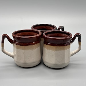 May include: Three ceramic mugs with a brown rim and interior, a tan band, and a speckled white base. Each mug has a handle with a tan accent. The mugs are arranged in a group.