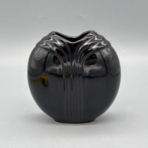 May include: A glossy black vase with a rounded body and a unique, stylized top. The vase has a smooth, reflective surface and a small, circular base. Vertical lines adorn the front of the vase, adding a decorative element. The vase is a decorative item.