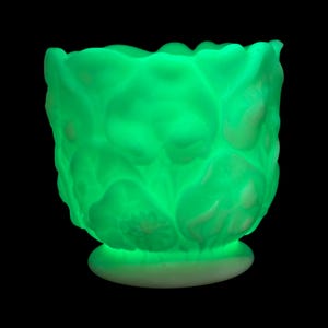 May include: A luminous green glass vase with a floral design. The vase features a scalloped edge and a circular base. The glass has a textured appearance, with light creating highlights and shadows. The background is black.