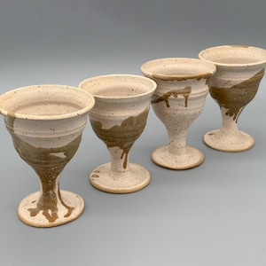 Puede incluir: Cuatro copas de cerámica hechas a mano. Cada copa presenta una base moteada de color blanquecino con un diseño de esmalte marrón goteado alrededor del borde y el tallo. Las copas tienen un cuenco ancho y redondeado y un tallo delgado.