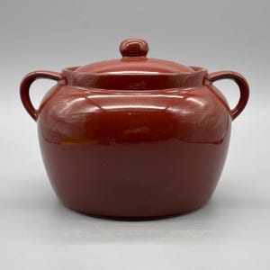 Könnte beinhalten: Ein runder, rotbrauner Keramik-Auflauf mit passendem Deckel. Die Schüssel hat zwei gebogene Griffe und einen kleinen Knopf auf dem Deckel. Die glänzende Oberfläche reflektiert das Licht. Ein klassischer Küchenartikel.