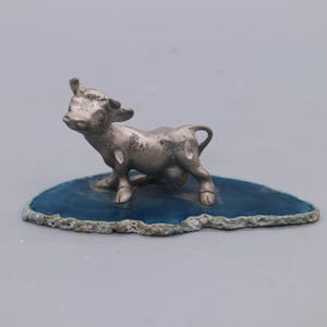 Op de afbeelding: Een kleine, zilverkleurige figuur van een stier staat op een blauwe, ovaalvormige basis. De stier heeft gedetailleerde kenmerken en staat. De basis heeft een getextureerde, natuurlijke rand.