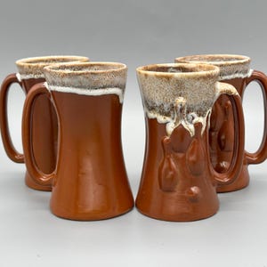 Puede incluir: Cuatro jarras de cerveza de cerámica marrón con un esmalte blanco y beige texturizado y goteado alrededor del borde. Cada taza tiene un asa grande y un diseño decorativo en relieve en la parte delantera. Las jarras están dispuestas en grupo.