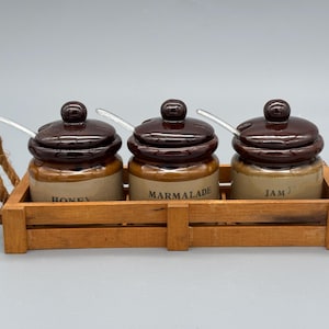 Könnte beinhalten: Ein Holztablett mit drei Keramikgläsern mit braunen Deckeln und Löffeln. Die Gläser sind mit "HONEY", "MARMALADE" und "JAM" beschriftet. Das Tablett hat Seilgriffe.