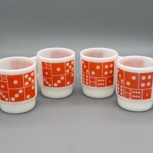 May include: Four white ceramic mugs with orange domino patterns. Each mug has a handle and a cylindrical shape. The domino patterns are in various arrangements, with white dots on an orange background. The mugs are arranged on a gray surface.