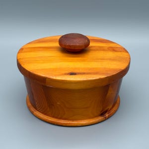 May include: A circular wooden container with a lid and a small, rounded knob. The container is made of light-coloured wood with a natural grain pattern. The lid and knob are a darker shade of brown.