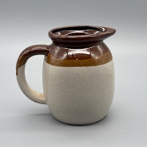May include: A ceramic jug with a brown rim and handle, a tan band, and a speckled off-white body. The jug has a spout and a curved handle. This kitchenware item is likely used for serving drinks.