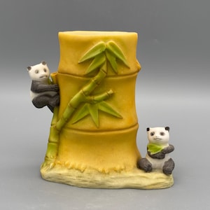 Puede incluir: Una figura de oso panda de cerámica con diseño de bambú. El jarrón amarillo con forma de bambú tiene hojas de bambú verdes y dos osos panda, uno trepando y otro sentado. Los pandas son negros, blancos y grises.